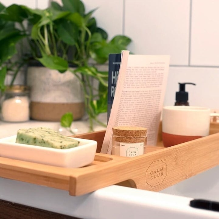 A wooden bath caddy with a book, wine, and other luxuries placed on it
