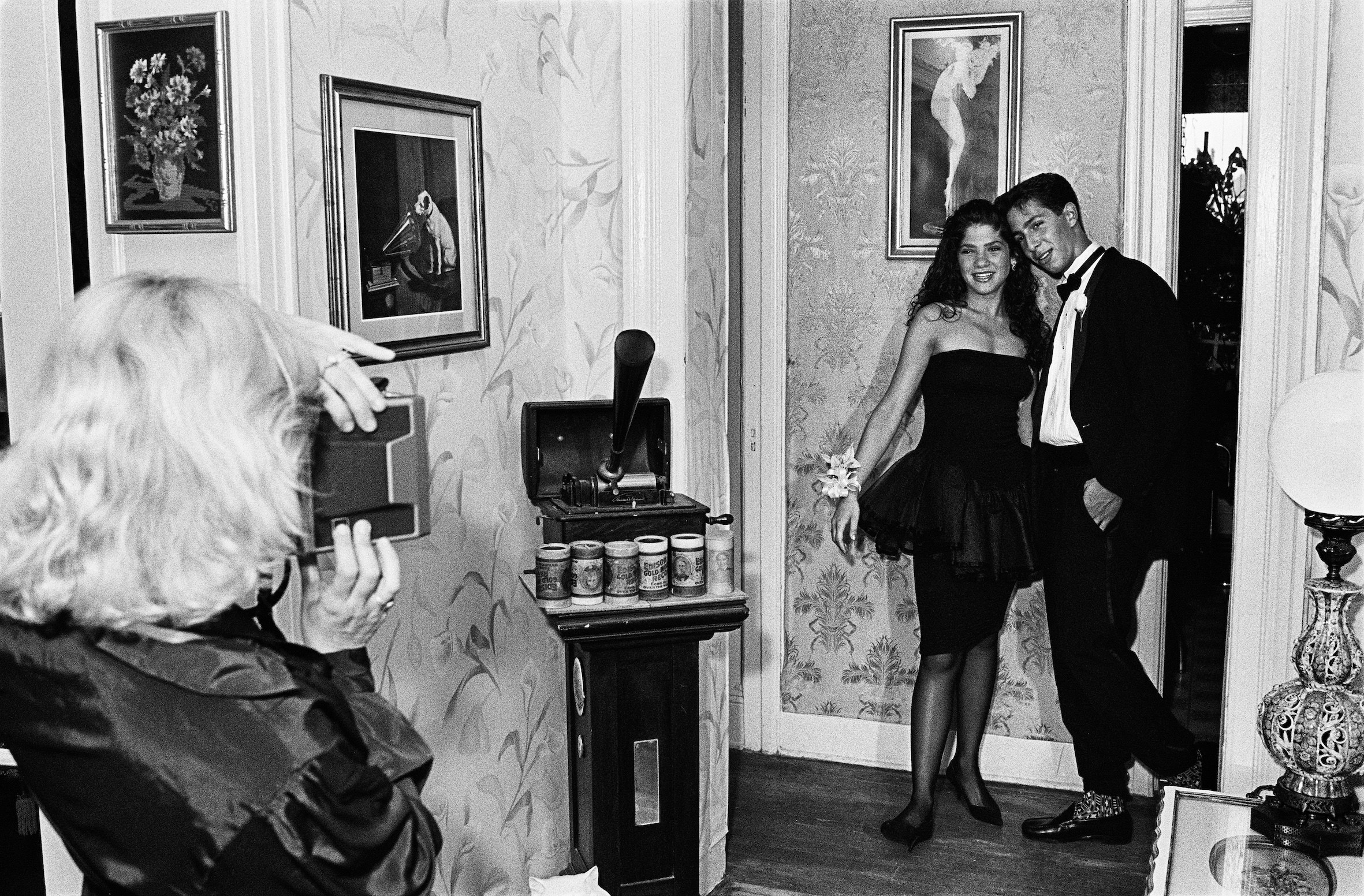 A woman takes a polaroid picture of a young girl and her prom date inside a house