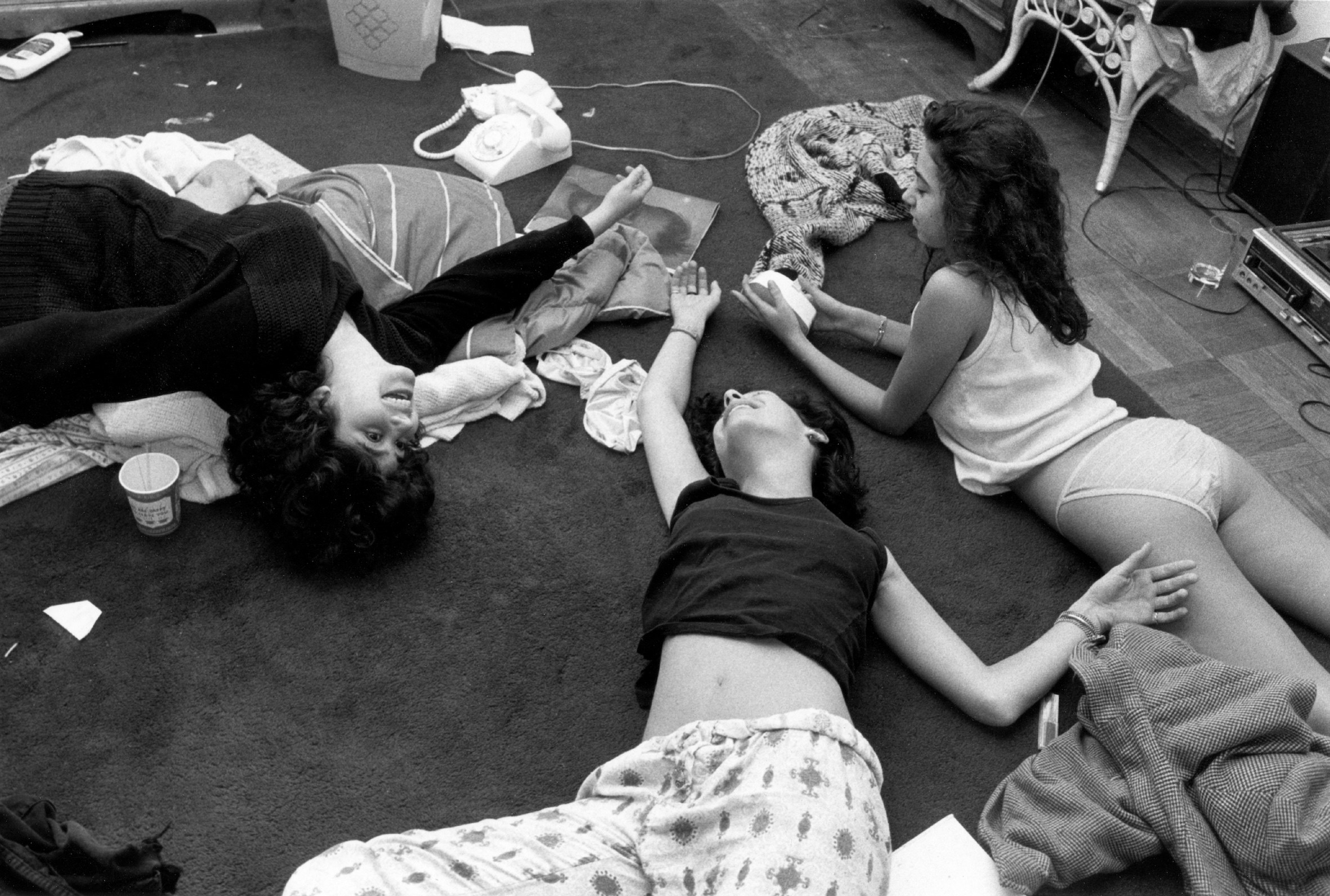 three girls lie on the floor in pajamas with phones, coffee cups, and some other items around them 