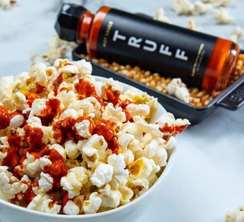 A bowl of popcorn topped with hot sauce and the hot sauce bottle in the background
