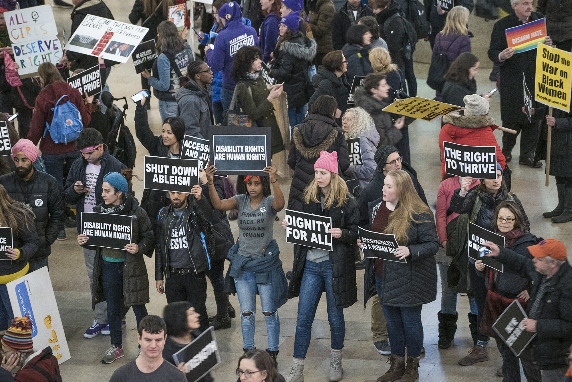 Congregated protesters carry signs that read &quot;shut down ableism&quot; and &quot;disability rights are human rights&quot; and &quot;dignity for all&quot;