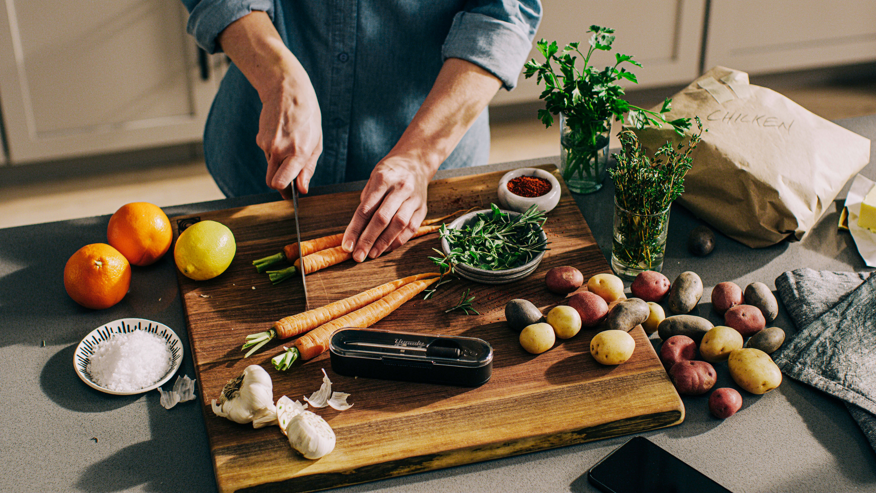 A person cuts carrots on a cutting board beside the Yummly® Smart Thermometer.