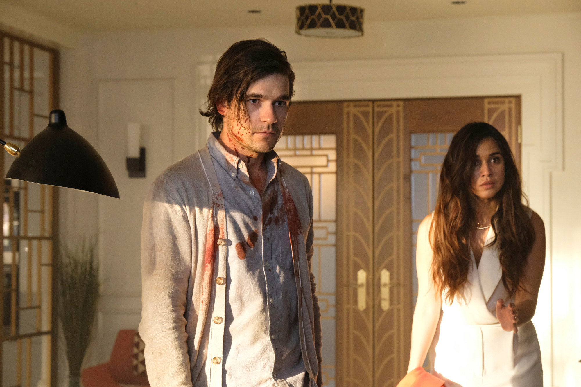 A man in a white shirt covered in blood stands looking at something calmly. A young woman behind him appears scared.