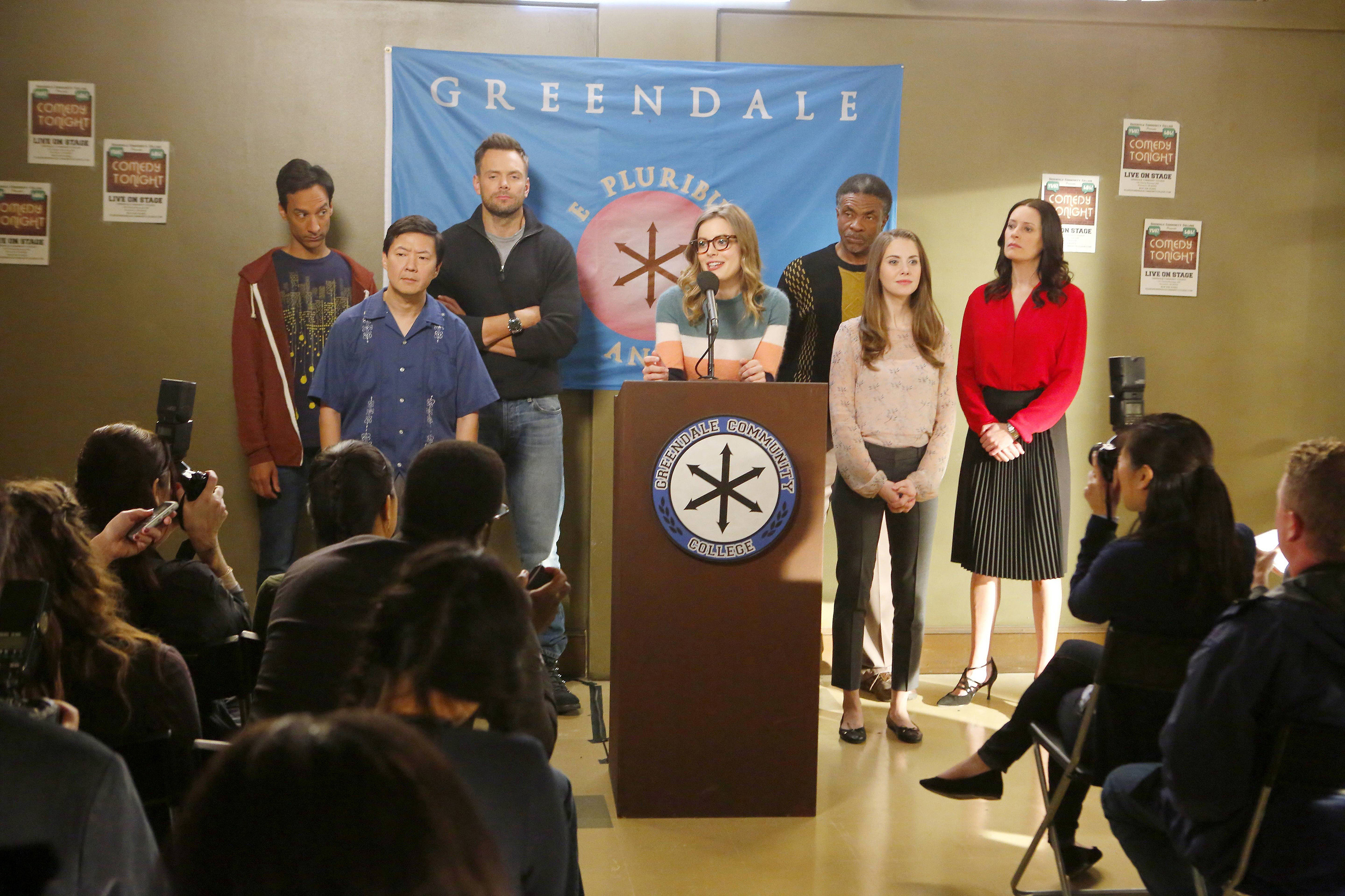 A group of seven community college students stand behind a podium, in front of a crowd. One woman talks into the podium&#x27;s microphone.