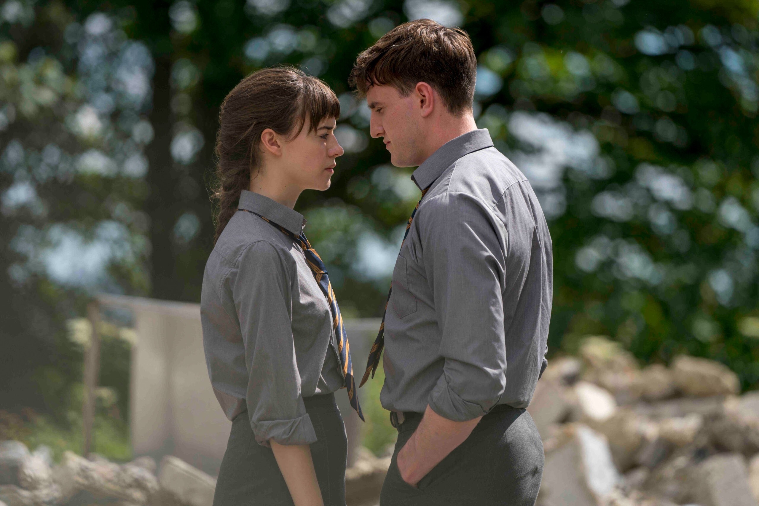 Two high school seniors, one boy on girl, stand in their grey school uniform, wearing ties. They stand very close to each other face to face with both longing and sad expressions.