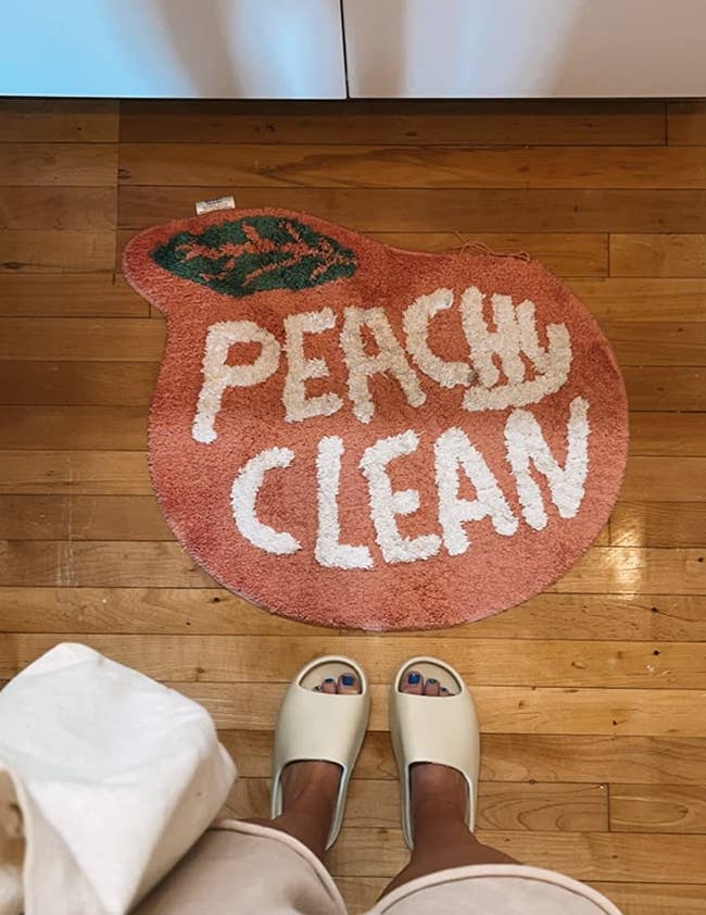 reviewer photo of them standing in front of the peach-shaped bath mat that reads 