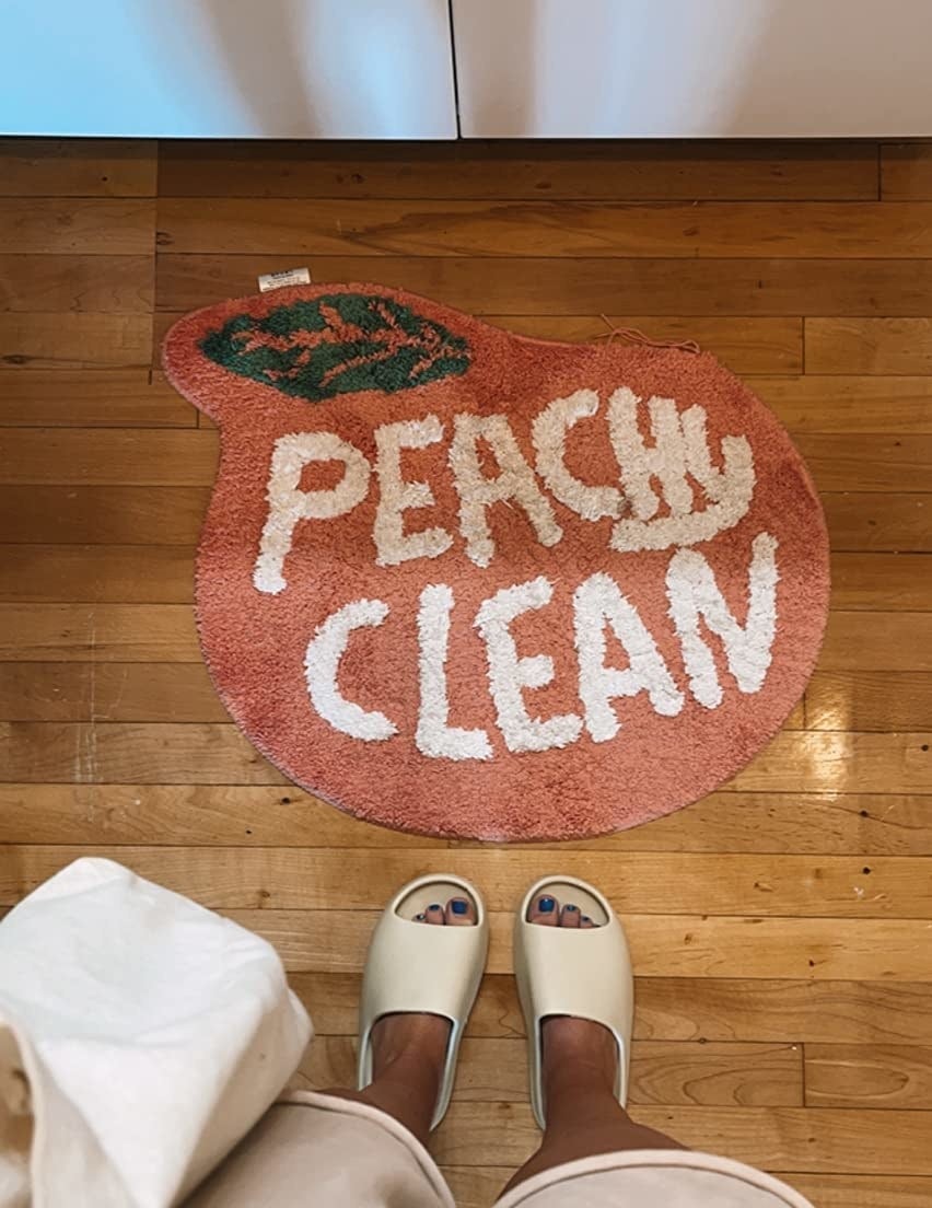 reviewer photo of them standing in front of the peach-shaped bath mat that reads 