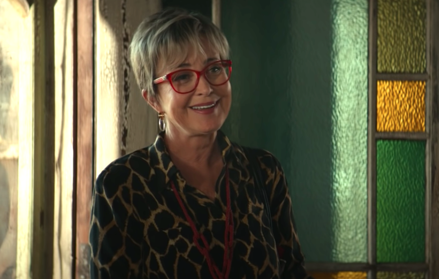 Annie Potts as Janine, standing in an old home with red framed glasses, smiling