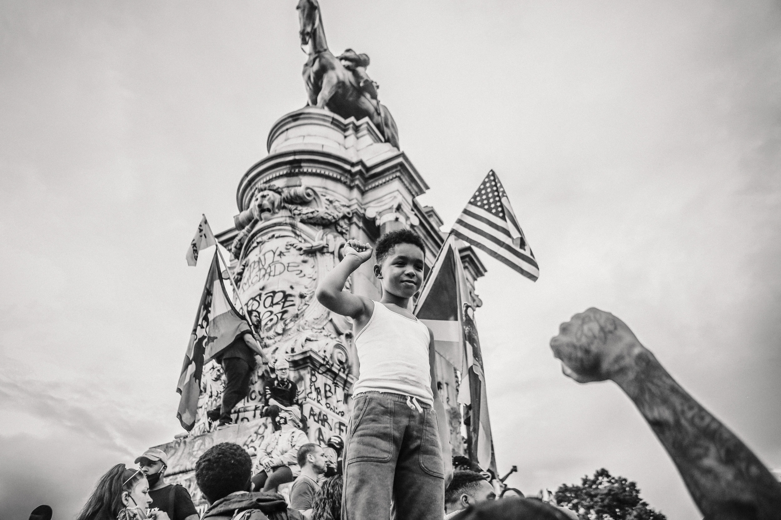 A young boy standing on a statue with his fist up and other protesters in the background with the American flag