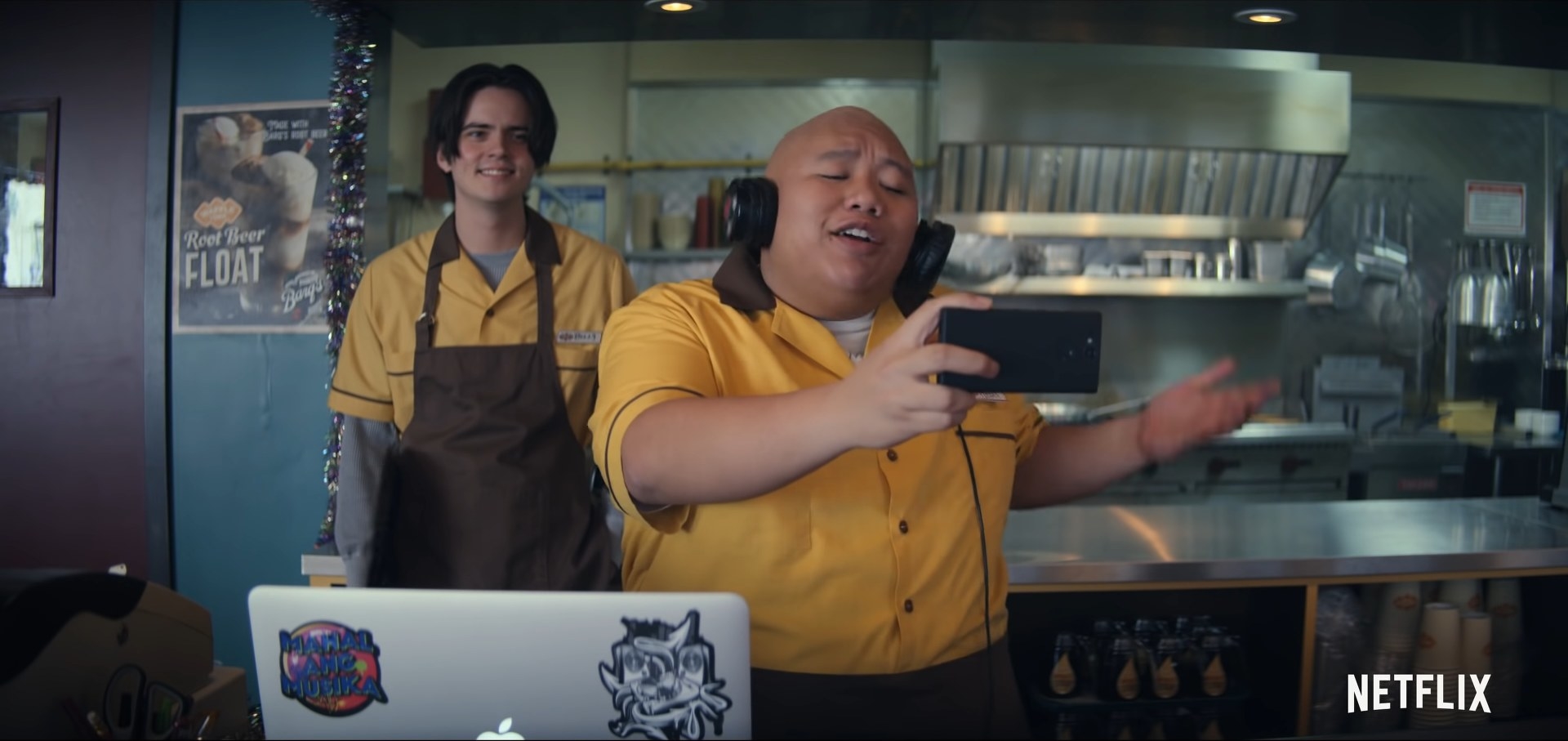 A restaurant worker wearing a pair of headphones, watches a music video on his smartphone while his co-worker looks at him from behind