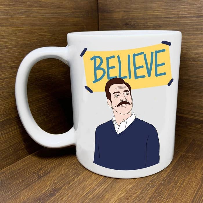 a white mug with ted lasso on it and a sign that says &quot;believe&quot; behind him