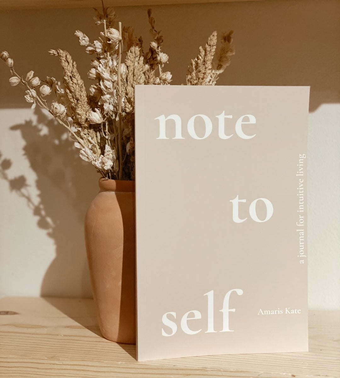 a tan journal leaning up a vase of flowers that says &quot;note to self; a journal for intuitive living&quot; on the cover