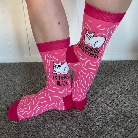 Person is wearing pink and red socks with a cat graphic and the words &quot;Cat hair on everything&quot; on one sock and &quot;Cat is the new black&quot; on the other