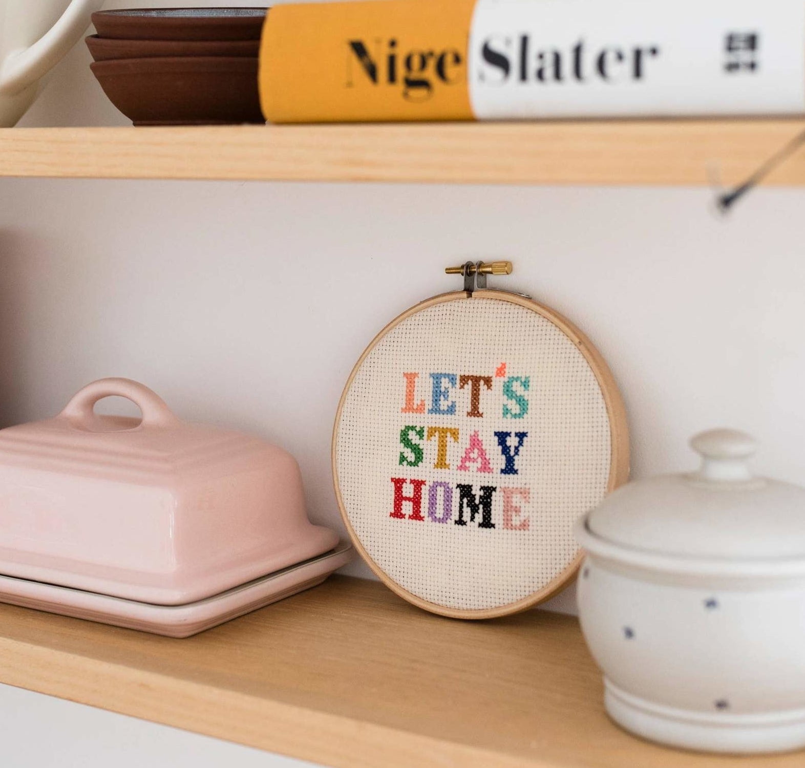 A cross stitch on a shelf that says &quot;Let&#x27;s Stay Home&quot; but each letter is a different color