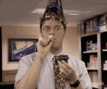 Dwight from the office wears a birthday hat and holds a cupcake with a candle in it