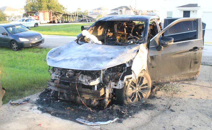 A destroyed car in a driveway is scorched black, surrounded by ash, with the windshield broken and front fender missing