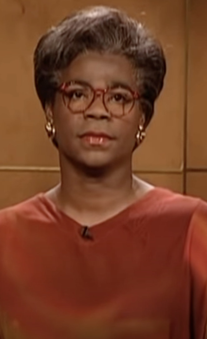 Morgan wearing an earth-toned blouse, glasses, lipstick, and earrings