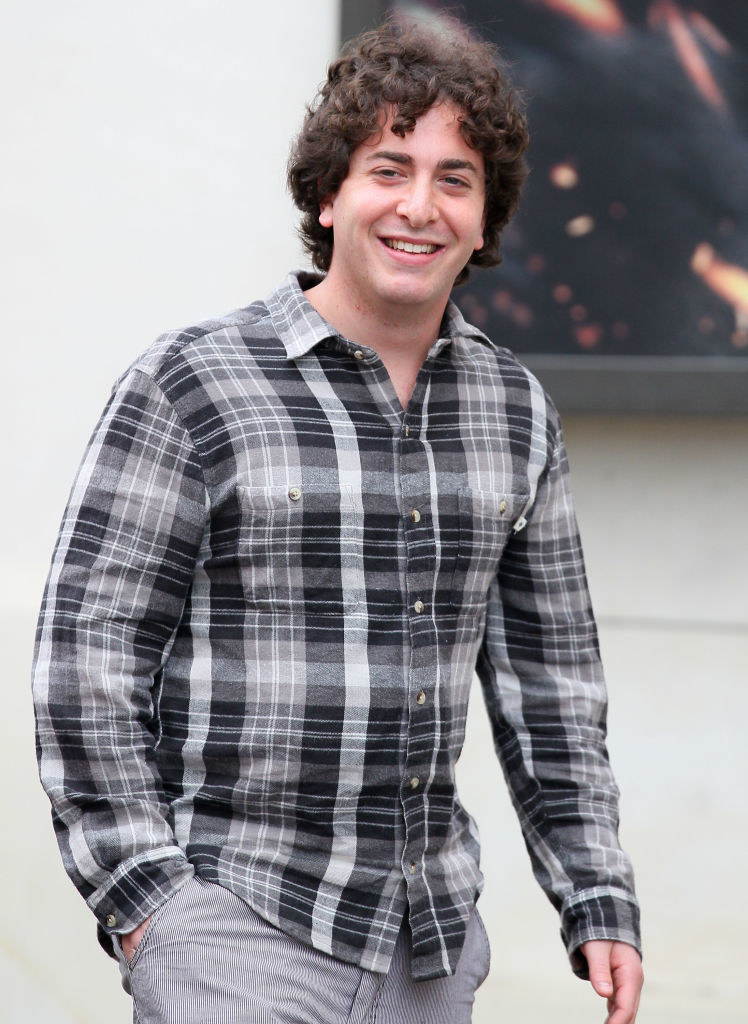 Man wearing a grey checkered shirt, and grey jeans. One hand in his pocket, mid walk smiling.