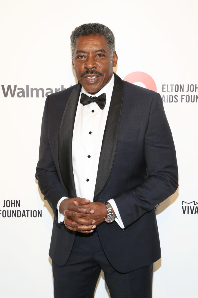 Man standing in a classic black suit, white shirt and bow tie. He&#x27;s clasping his hand together in front of him smiling.