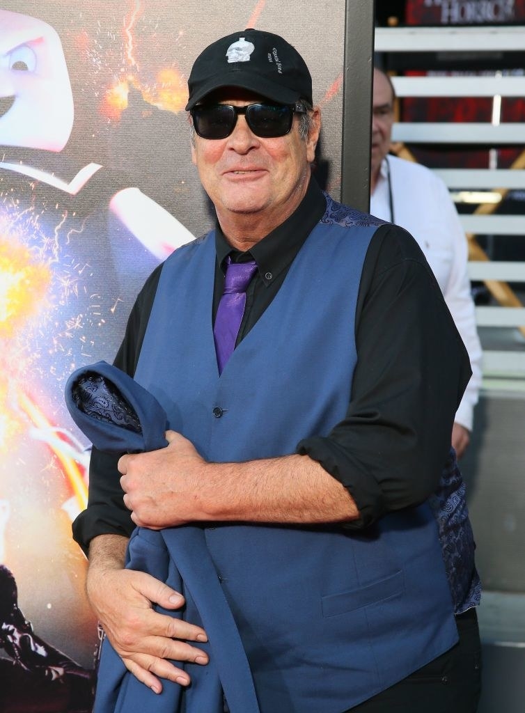 Man holding a blue blazer, wearing a matching blue vest and black shirt with a purple tie. His sleeves are rolled up and he&#x27;s wearing dark sunglasses and a black cap.