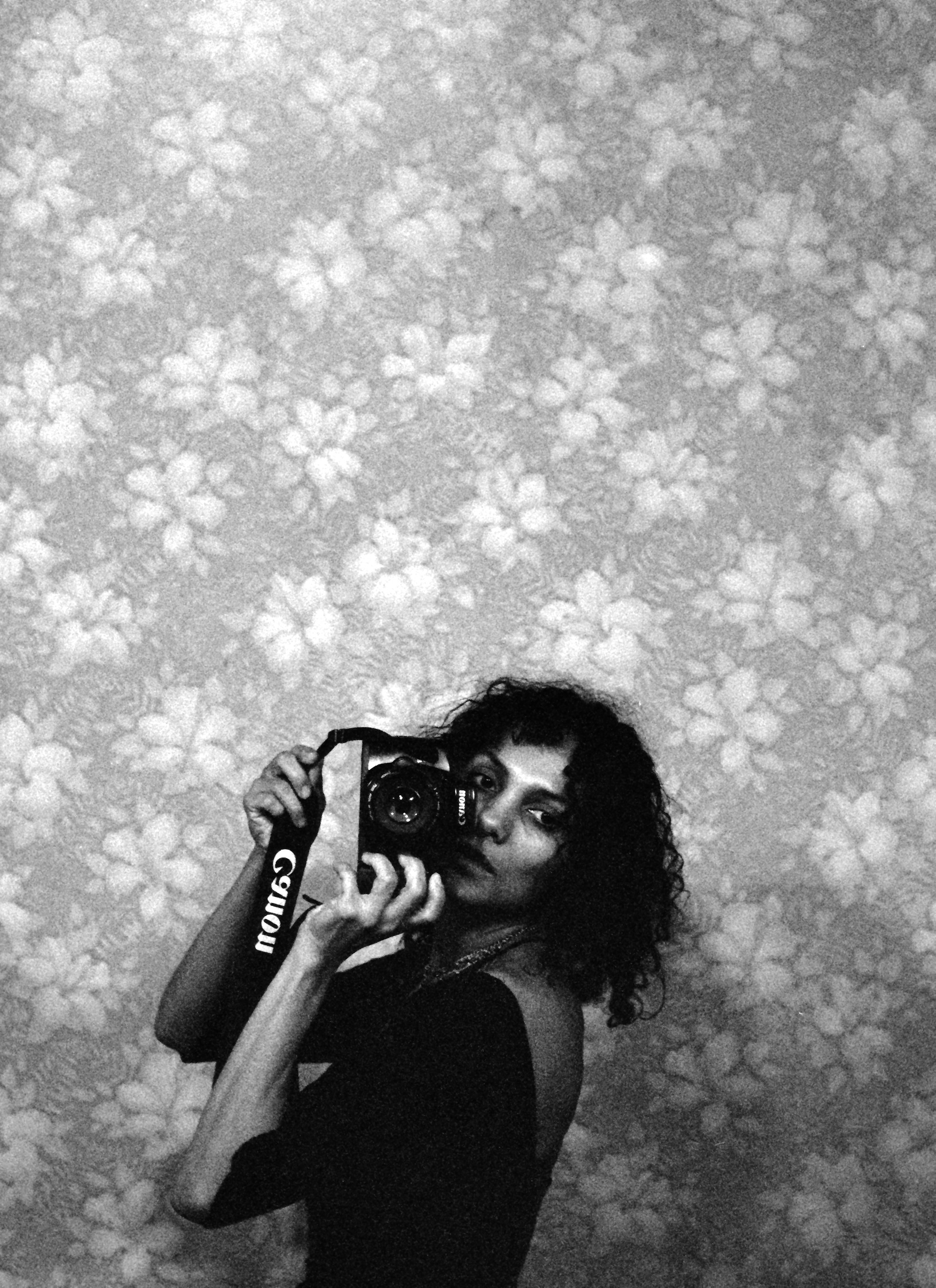 A self-portrait of the artist Ming Smith with a Canon camera in front of a floral wallpaper background