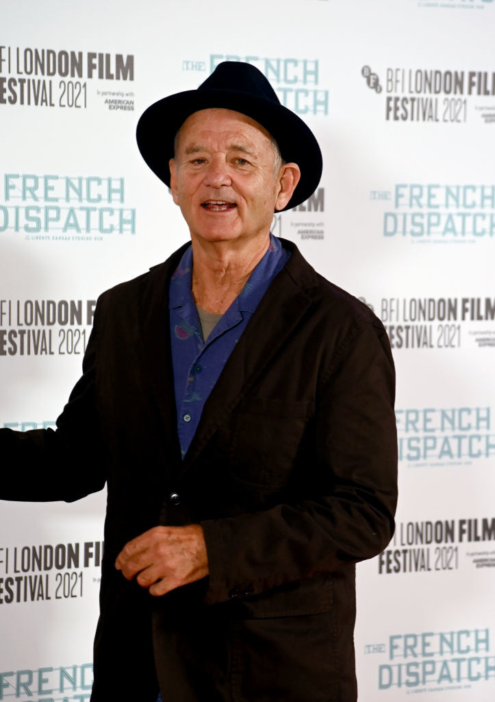 Older man wearing a black suit with a deep blue shirt underneath. He has a black hat on and he&#x27;s pointing off to the left smiling.
