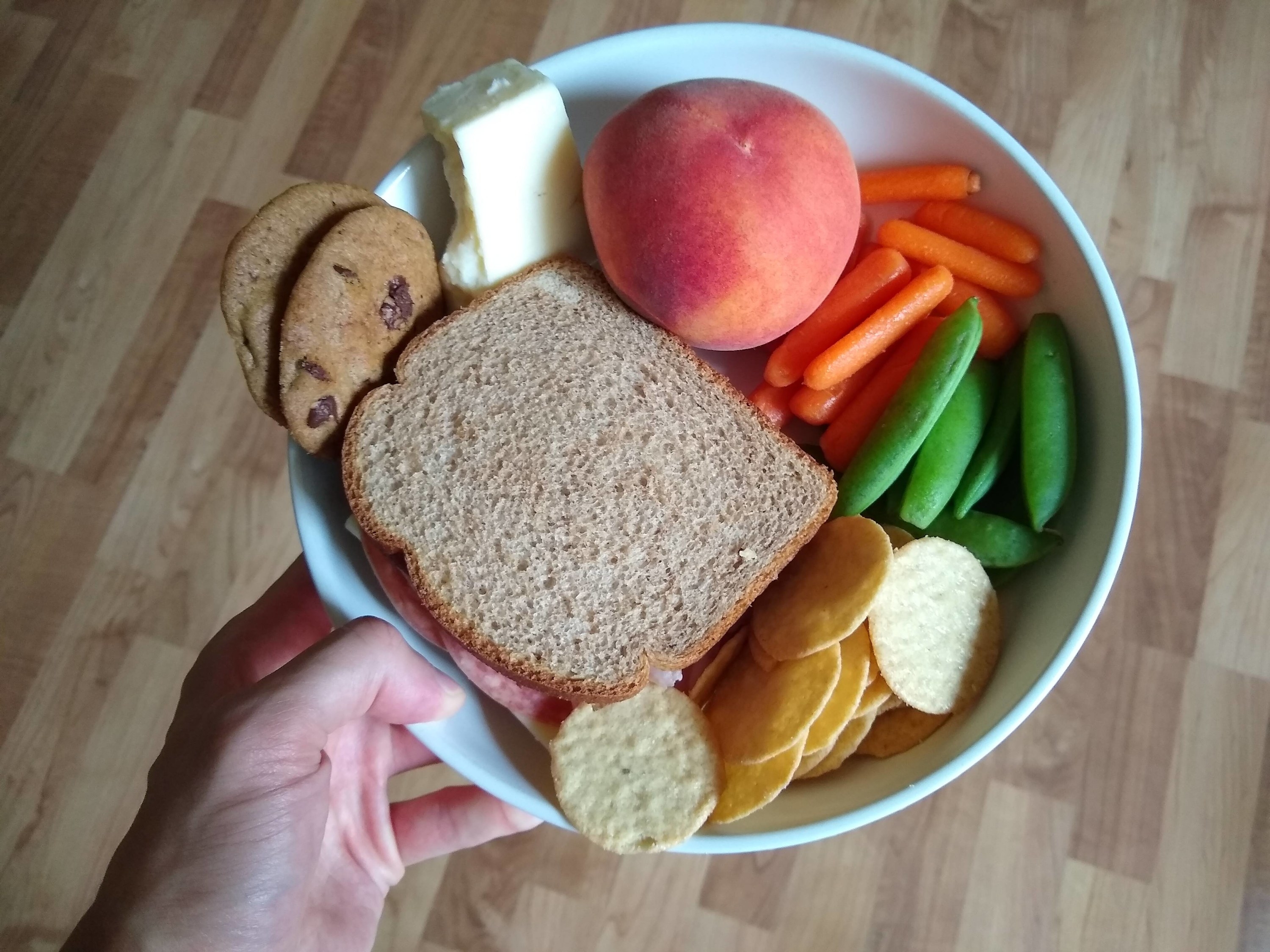 A bowl filled with random foods like bread, chips, carrots, cheese, cookies, and a peach.