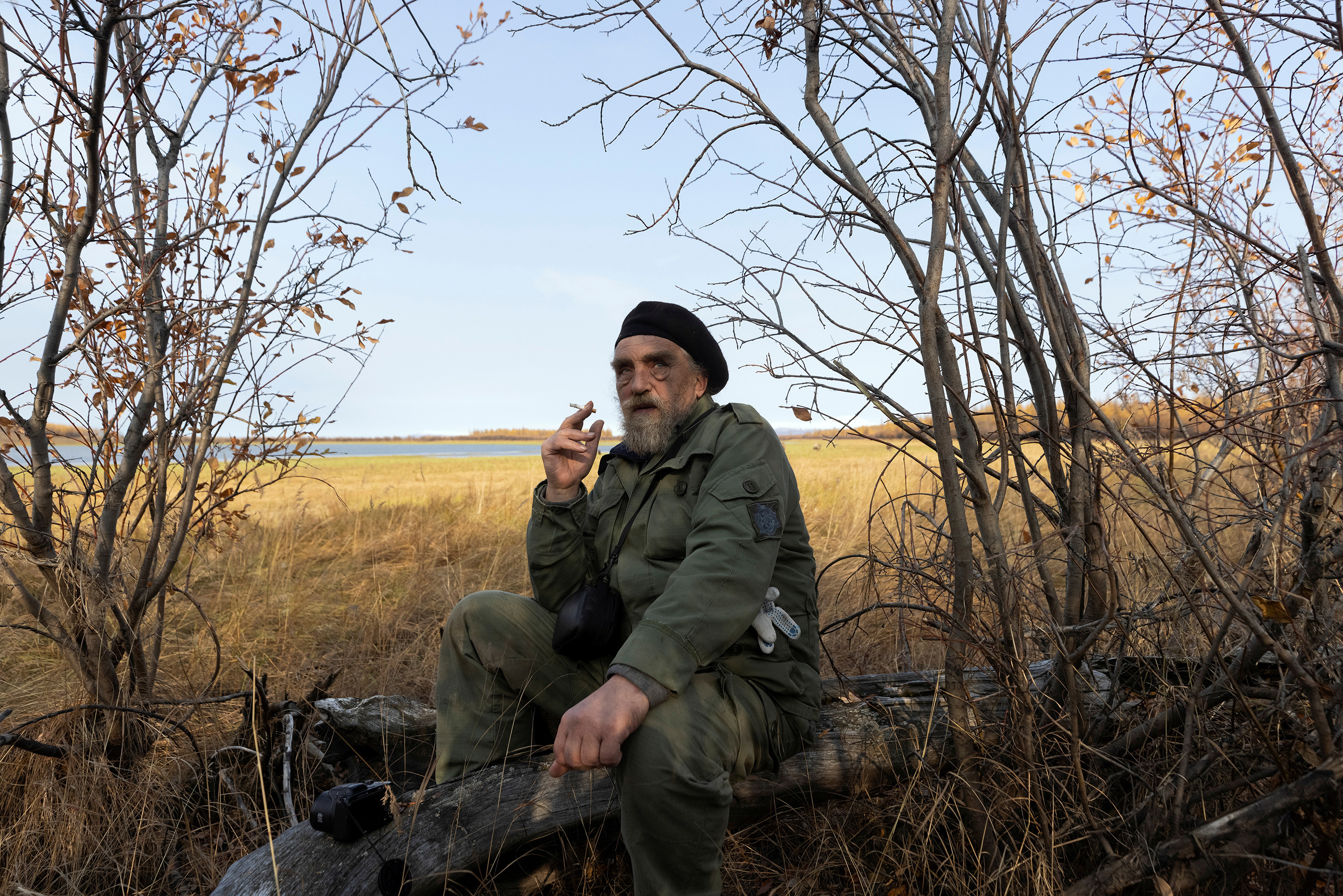 A man in an olive green jacket and pants sits on a fallen tree and smokes a cigarette