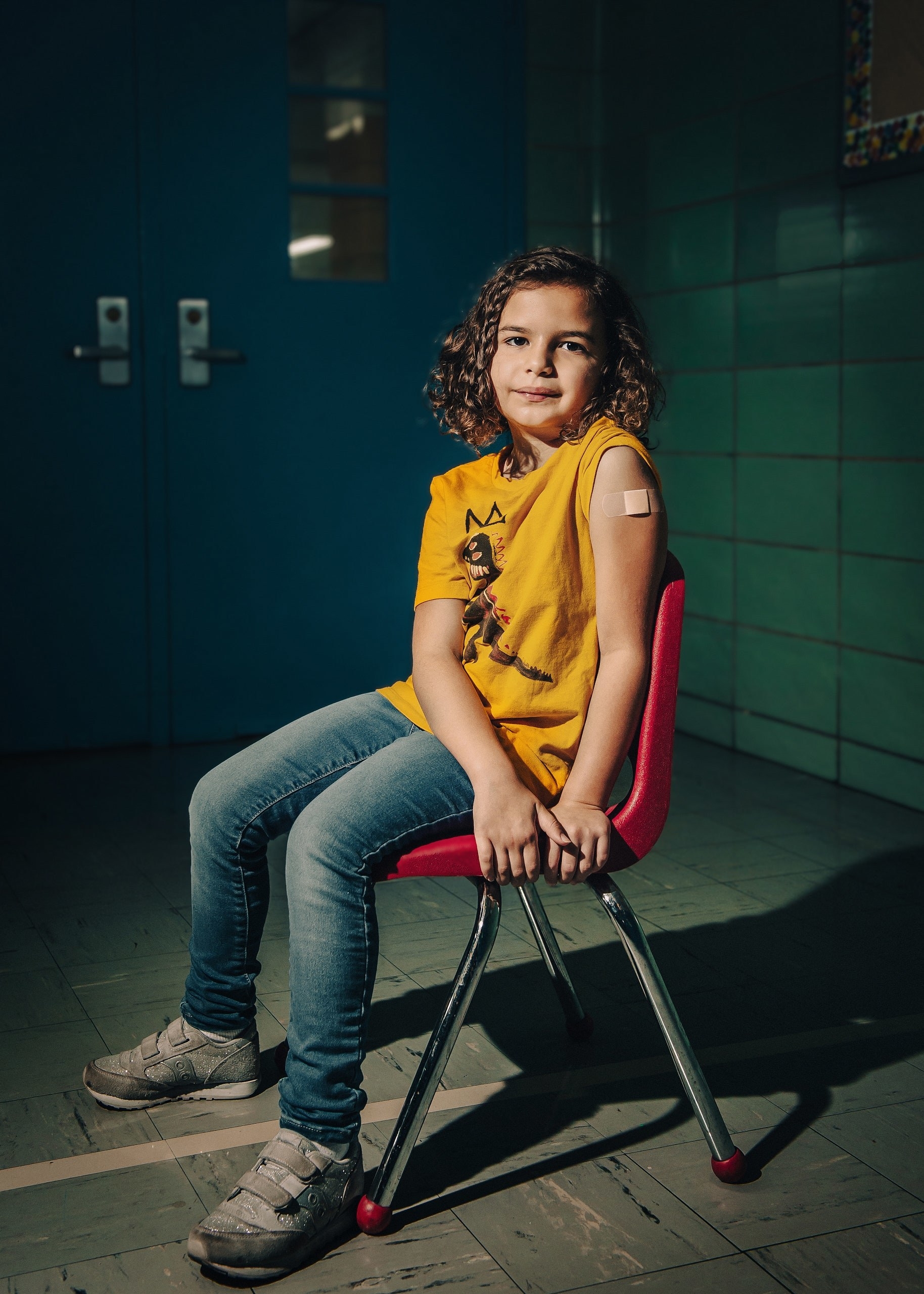 A little girl sits on a chair with a bandage on her left upper arm