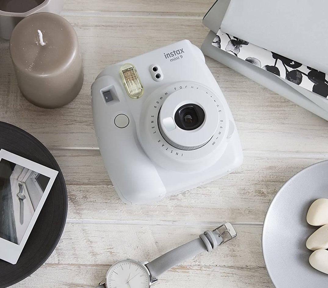 a white Fujufilm instax camera laying beside film, candles, and other desk decor