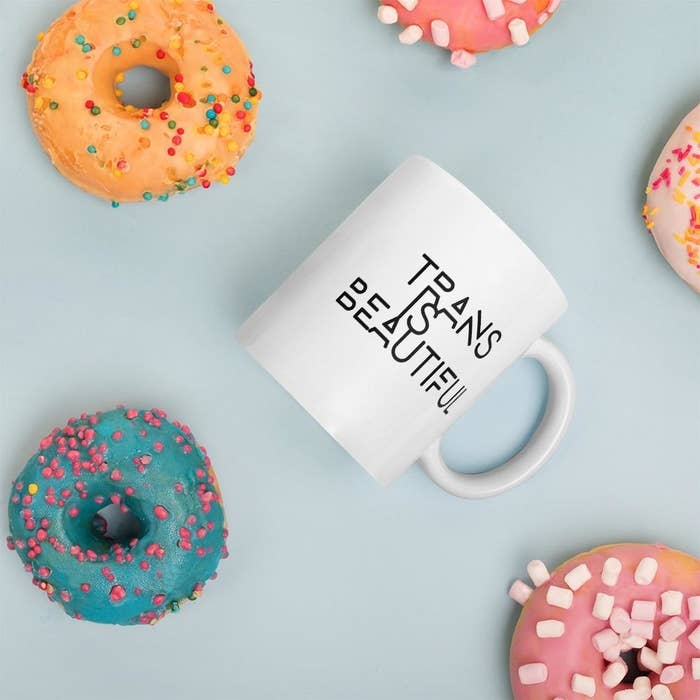 a white trans is beautiful mug with black lettering surrounded by doughnuts