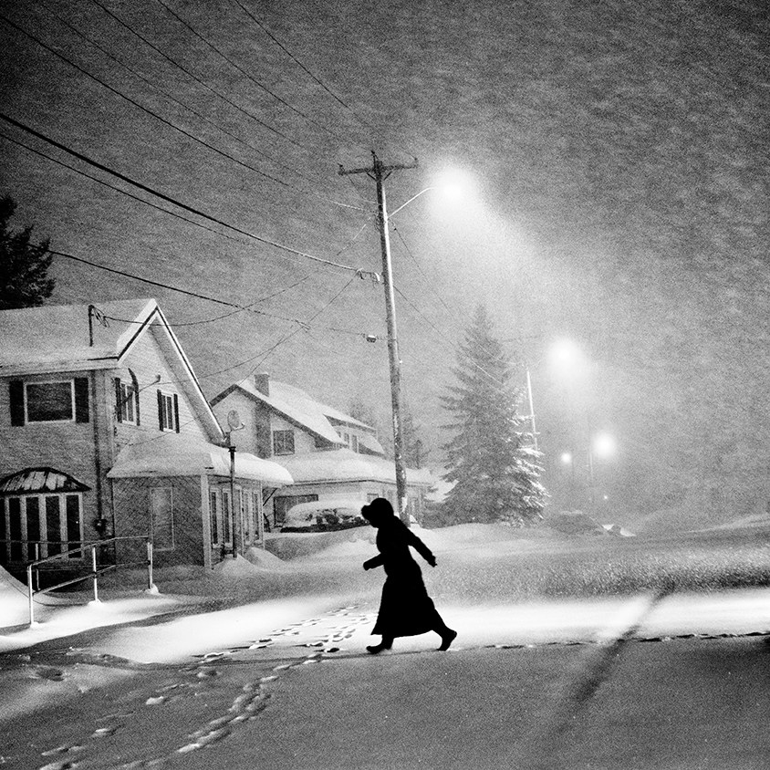 A person wearing a thick snow jacket walks through a snowy street in the middle of a storm