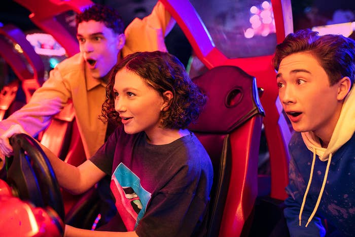 A woman playing a racing arcade game while two men watch.