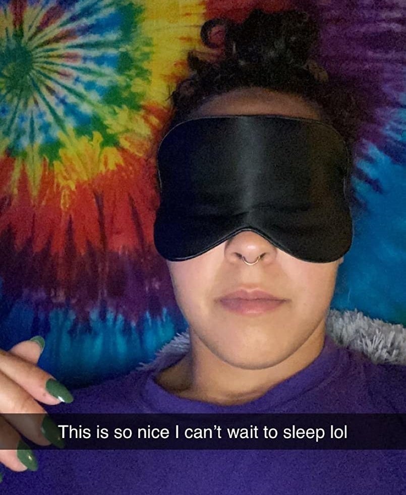 A reviewer wearing the eye mask in black with the SnapChat caption, &quot;This is so nice I can&#x27;t wait to sleep lol&quot;