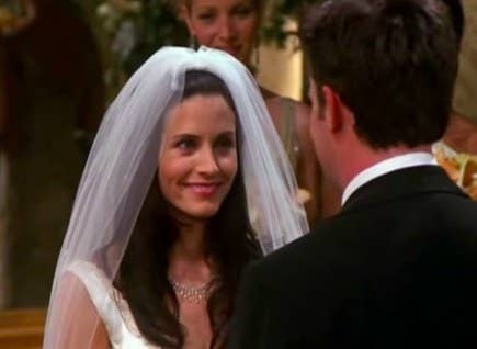 Monica wears a wedding dress and veil and smiles up at Chandler, who wears a suit