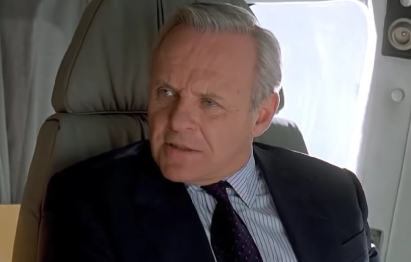William sits in an airplane seat wearing a suit and looking to the side as he speaks