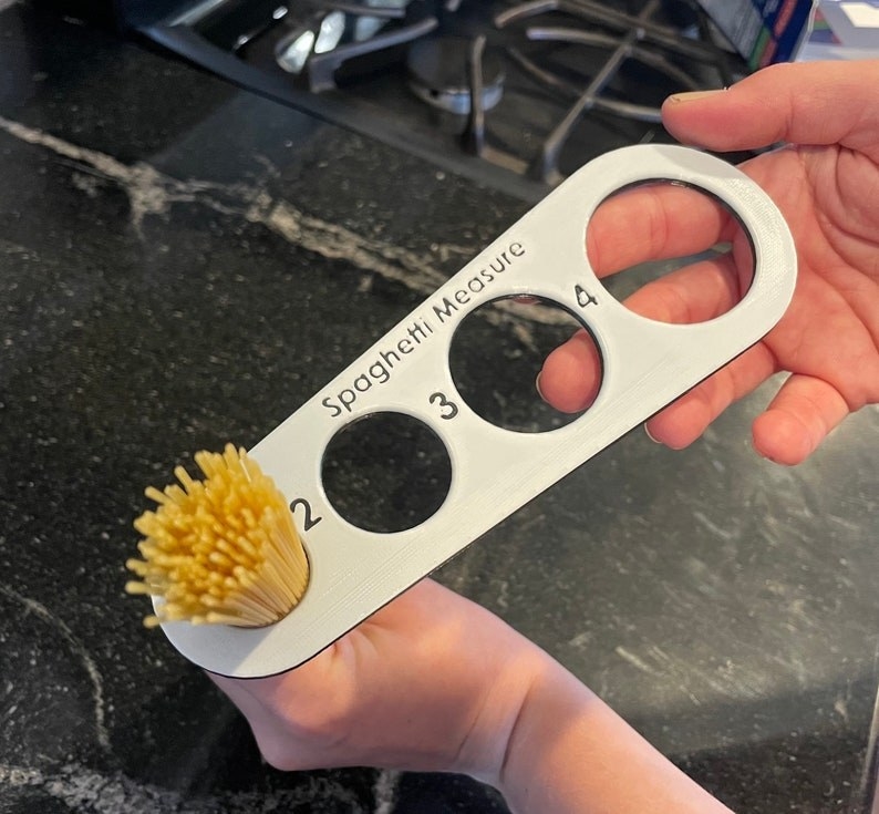 hands hold silver measuring tool with different-sized slots for dry spaghetti