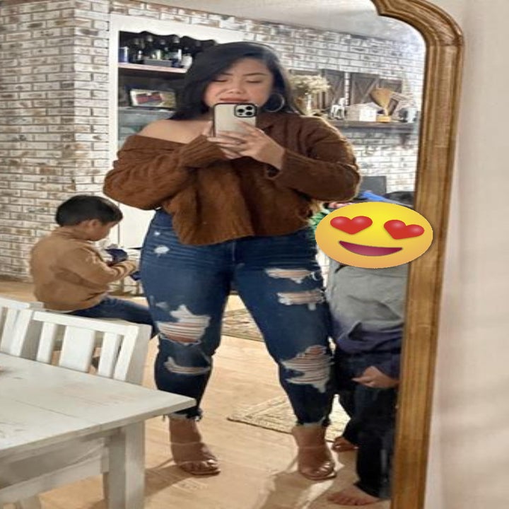 A reviewer's mirror photo while wearing the brown hoodie sweater in an off-the-shoulder style with distressed jeans and heels