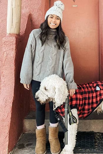 reviewer photo of them wearing a gray turtleneck sweater with a white pompom hat, black leggings, and brown boots while standing next to their dog