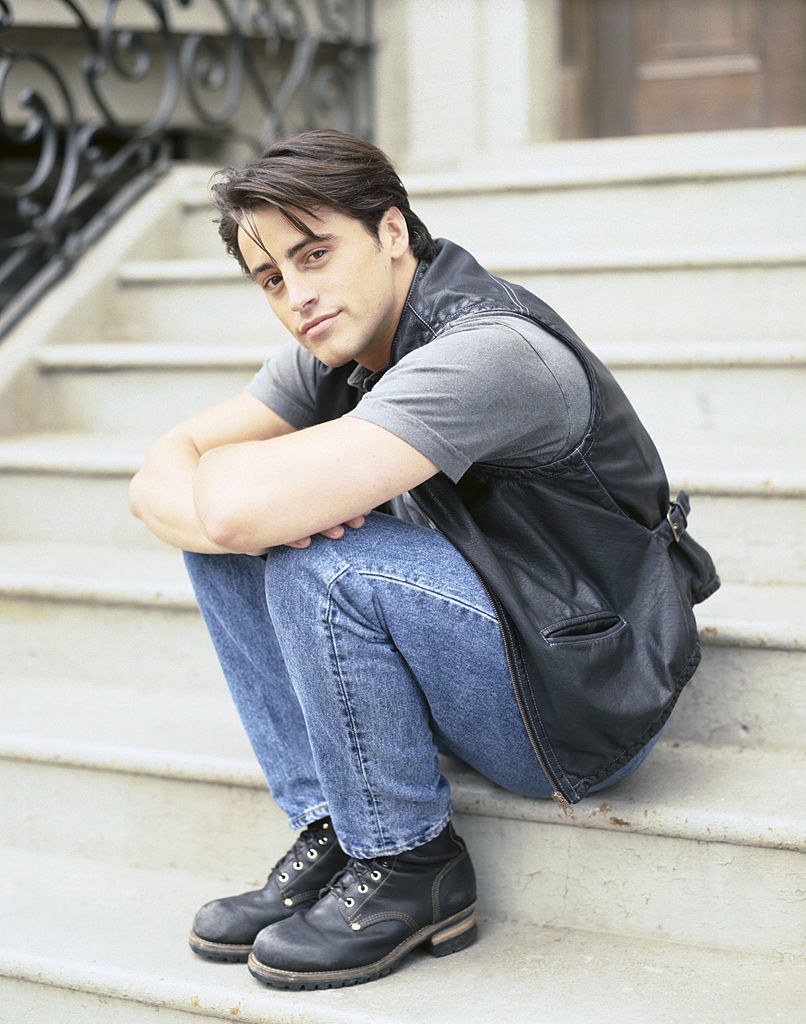 Joey wearing jeans, a shirt, and a leather-looking vest and sitting on steps