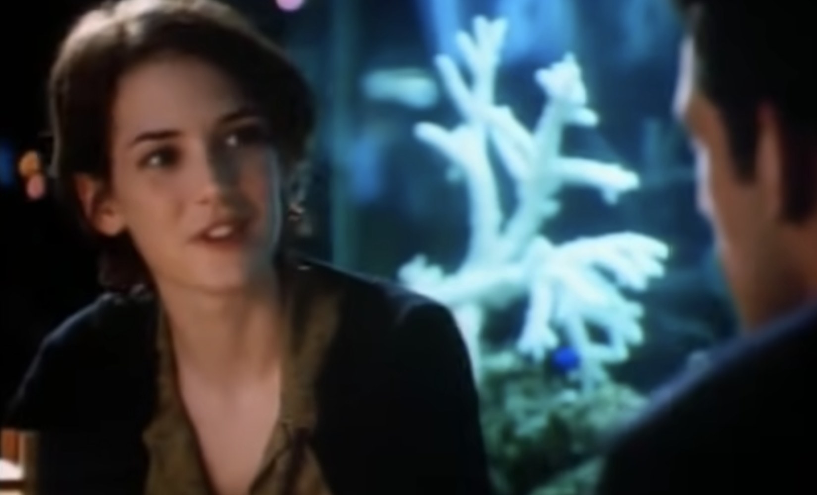 Winona Ryder smiles with her head cocked sideways. Behind her is a fishtank with coral.