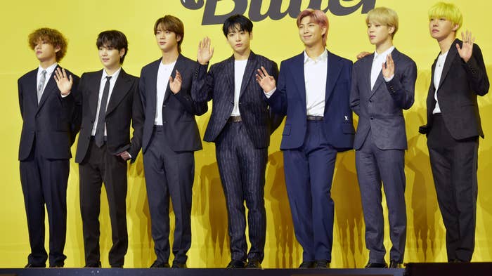 The members of BTS stand together in a line, most of them waving