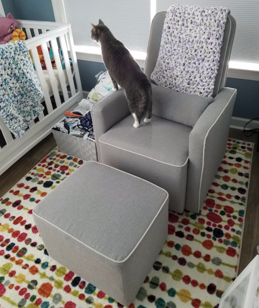the gray glider and ottoman with a cat on it next to a crib
