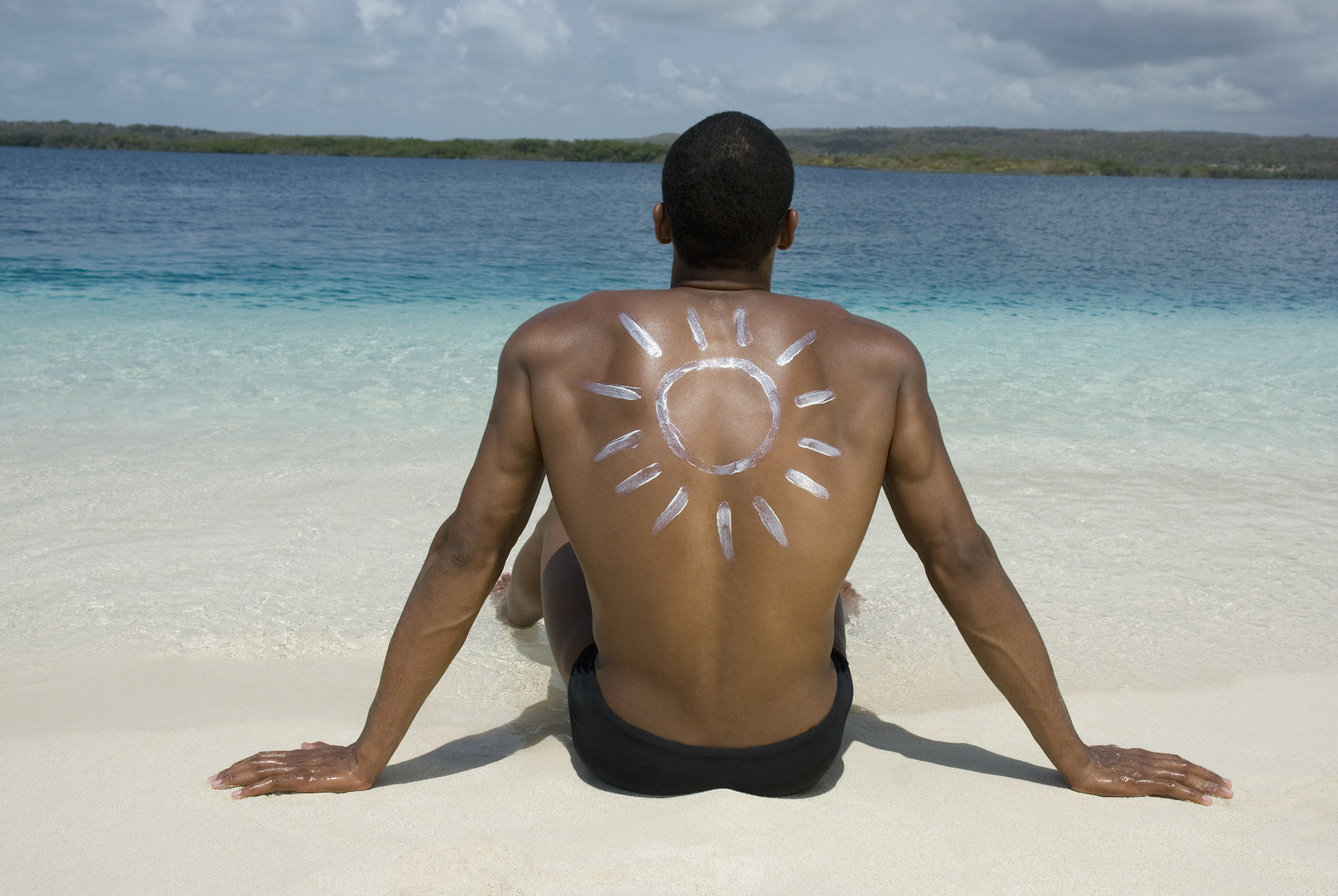 A guy sitting on the beach with a picture of the sun painted on his back with sunscreen