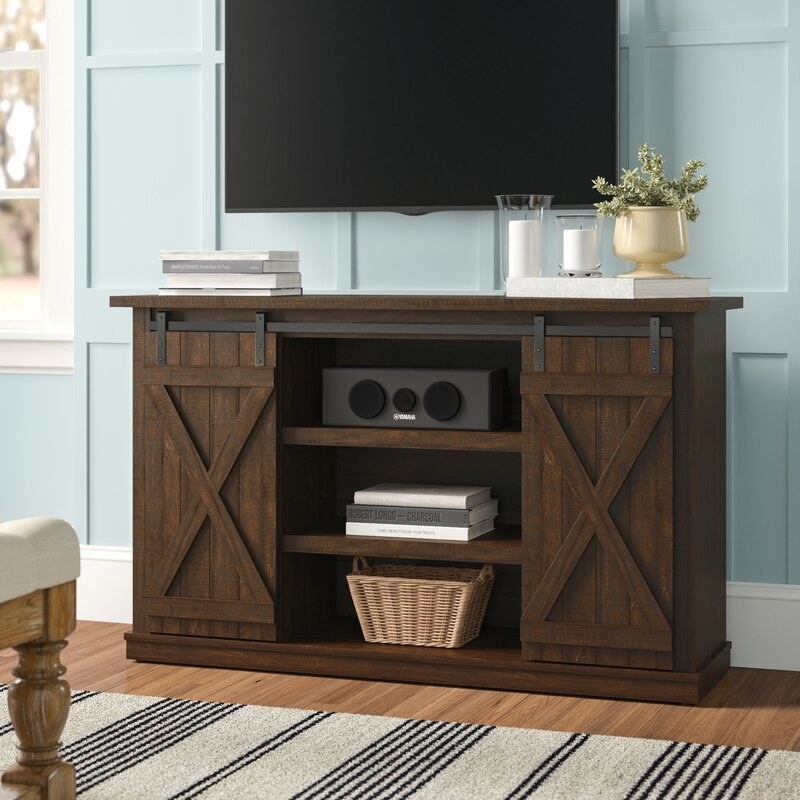 Brown farmhouse style TV stand, barn doors open to reveal two inner shelves with console, books, and basket, books, candles and vase on top, TV above stand
