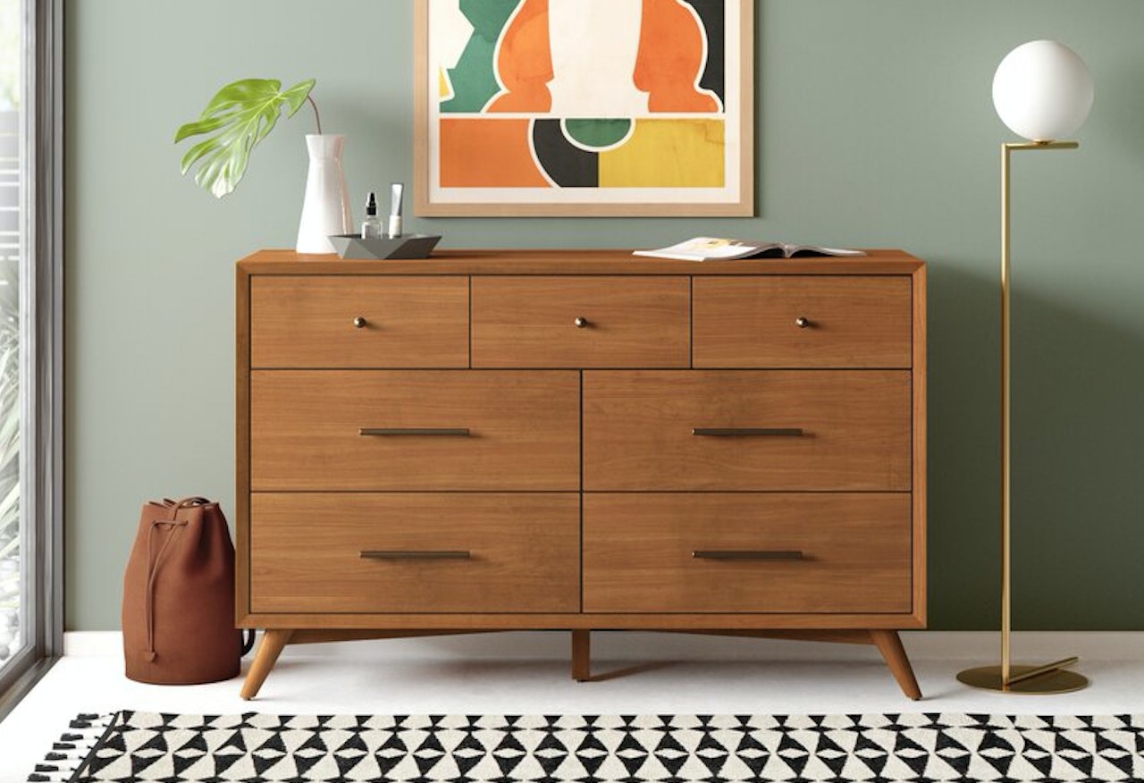 Acorn colored wooden dresser with seven drawers, vase, dish, book on top, geometric picture behind dresser, gold floor lamp on right side, brown backpack on left side floor