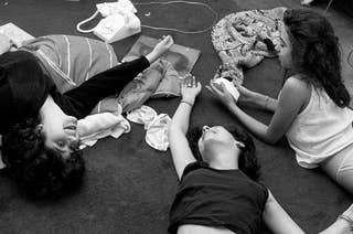 three girls lie on the floor in pajamas with phones, coffee cups, and some other items around them 