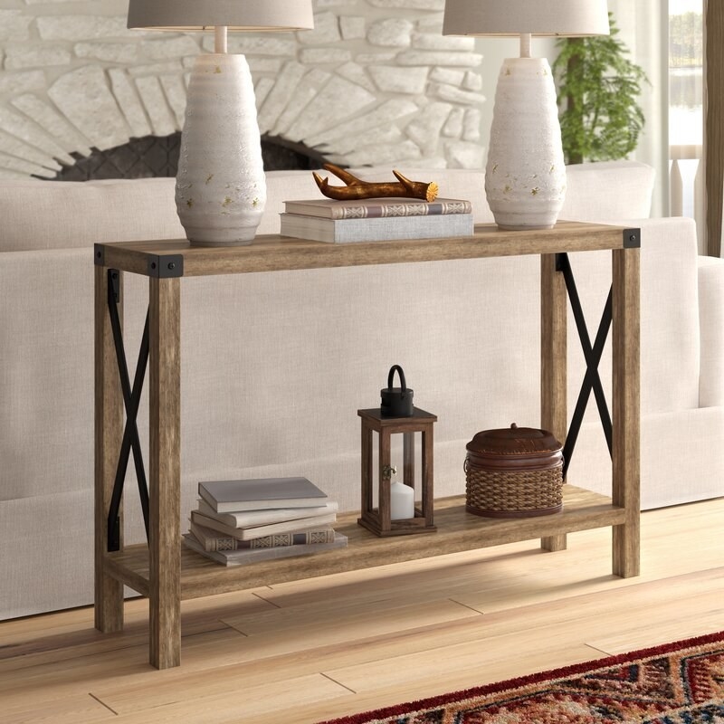 Wooden console table with books, candle holder, and basket on bottom shelf, books and two lamps on tabletop