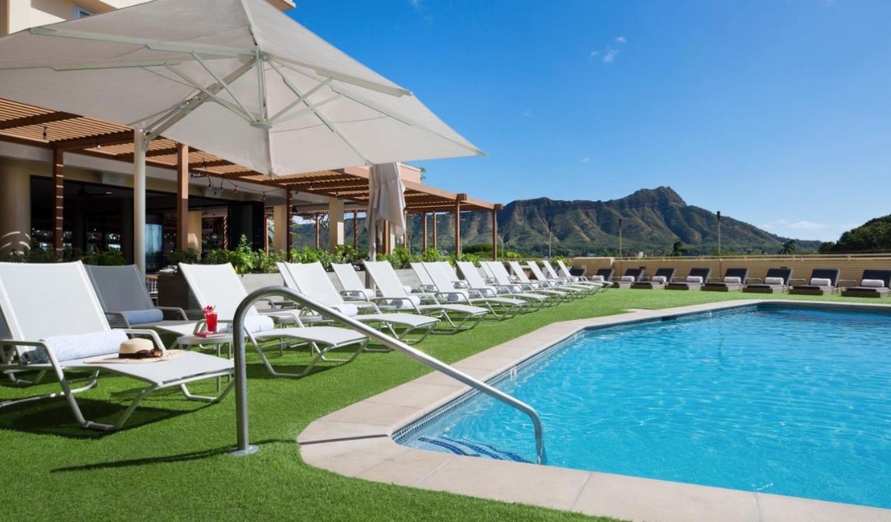 A photo of the pool at the Queen Kapiolani hotel, with grass along the edge and a row of loungers
