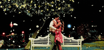 Rafael lifts Jane as they kiss in front of a tree and bench with petals falling around them